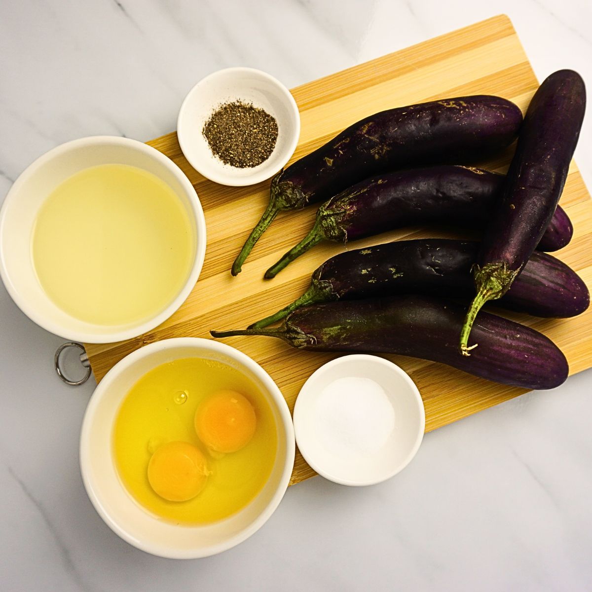 Bowl of oil, eggs, salt, pepper and eggplants on a wooden board.