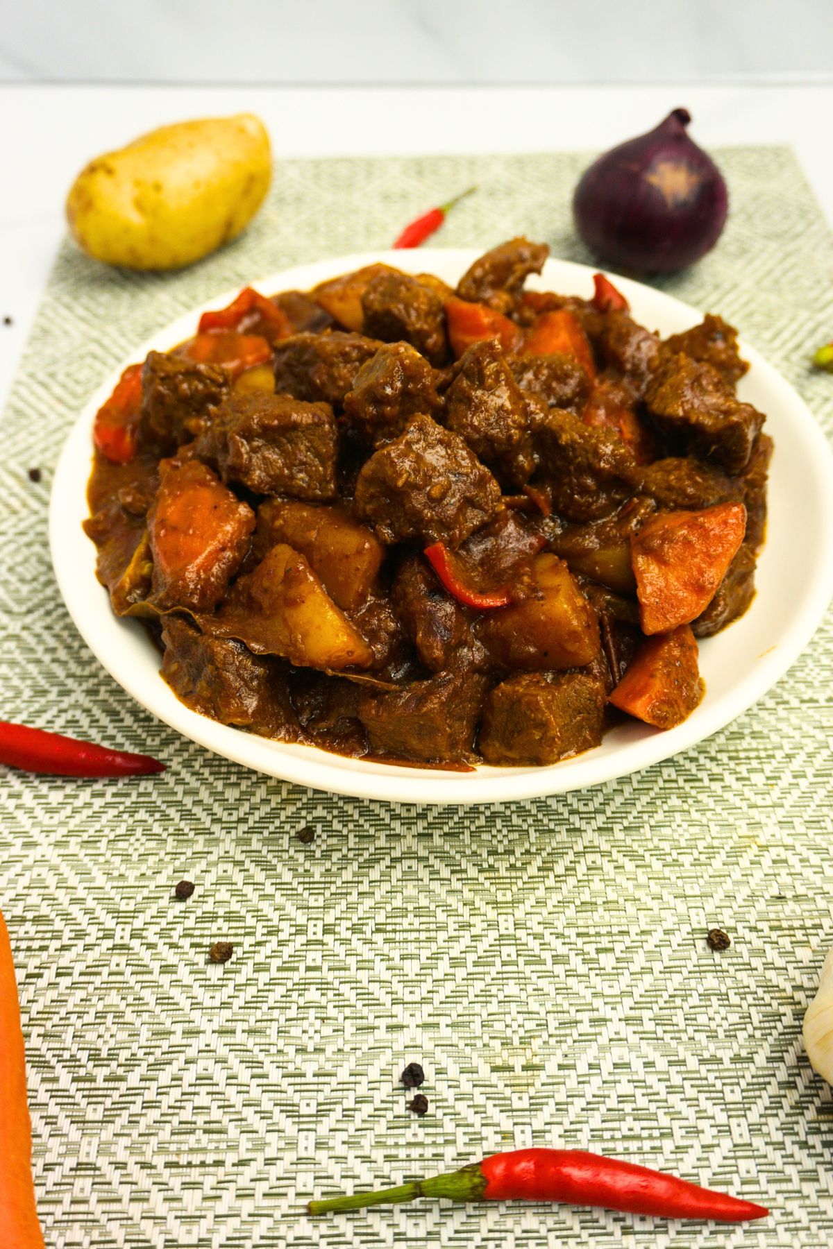 Spicy beef stew in a plate with fresh onion, potatoes, carrots, chilis and black pepper on a table.