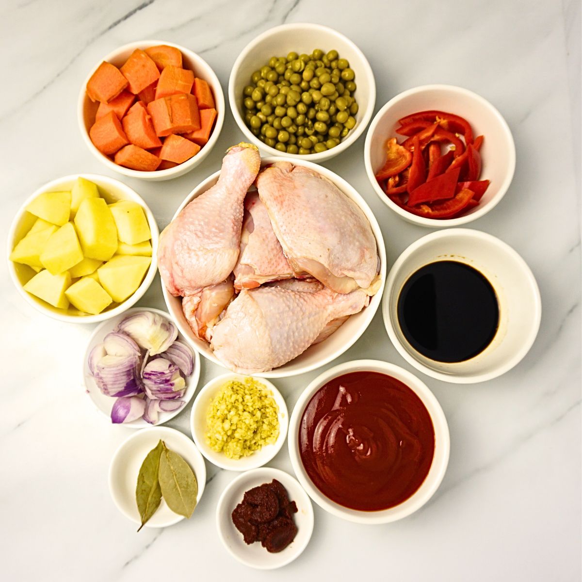 Chicken afritada inggredients in bowls. chicken, onion, garlic, bay leaves, potatoes, carrots, green peas, bell pepper, soy sauce, tomato paste and tomato sauce.
