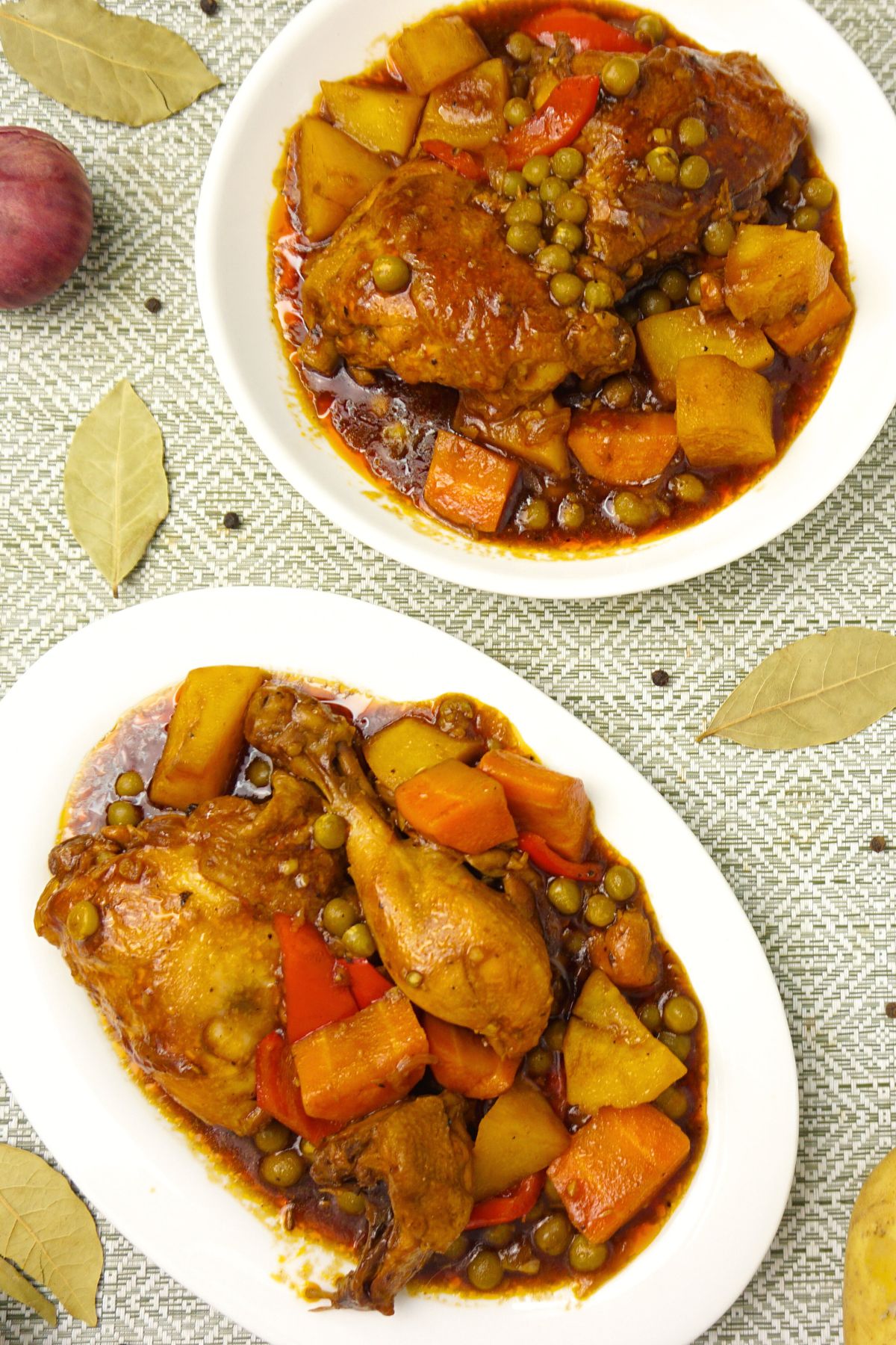 Top view of two plates of chicken afritada on a table with fresh bay leaves, onion and black pepper.